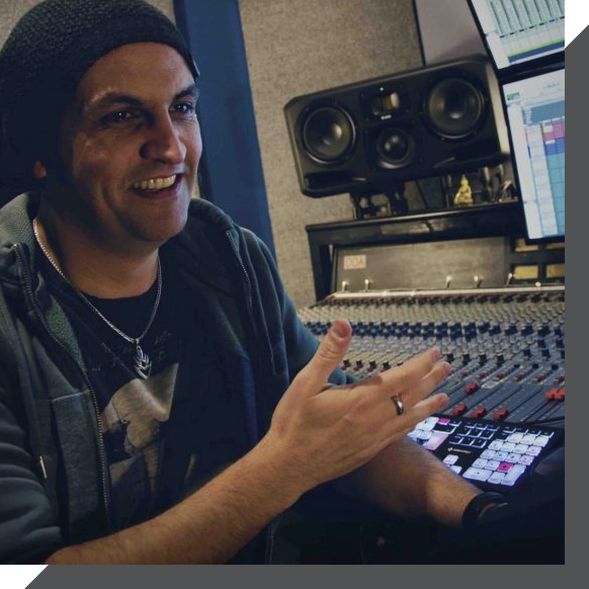 A person wearing a beanie and a dark jacket is sitting in a recording studio, gesturing with one hand while the other rests on a control panel with illuminated buttons. Large speakers and a monitor displaying audio levels are visible in the background.