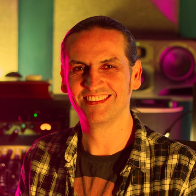 A person with long hair tied back is smiling in a room with various audio equipment. The individual is wearing a plaid shirt over a graphic t-shirt and is illuminated by colored lighting.
