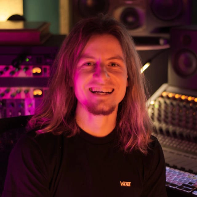A person with long hair and a goatee, wearing a black Vans t-shirt, smiles while sitting in a room filled with audio equipment. The lighting in the room has a pinkish hue.