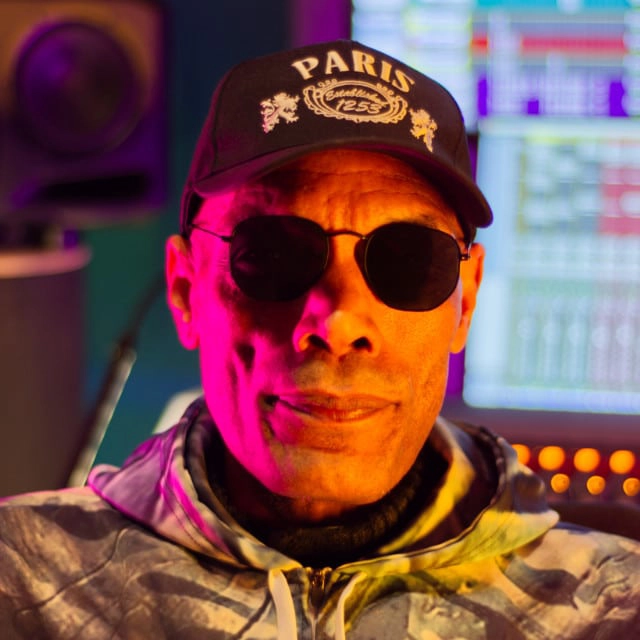 A person wearing a Paris cap and sunglasses is pictured in a recording studio. The background includes audio equipment and a computer screen displaying various audio levels and controls. The lighting casts a pink hue on the side of the person's face.