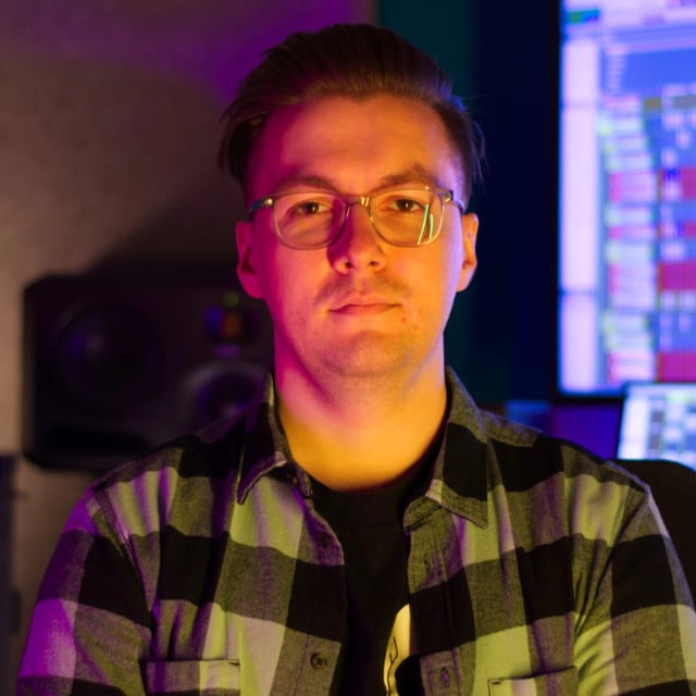 A person wearing glasses and a black-and-white checkered shirt is sitting in a room with audio equipment in the background. The room is lit with colorful lighting, creating a vibrant atmosphere.