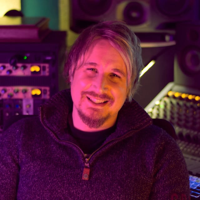 A person with medium-length hair and a beard is smiling at the camera. They are wearing a dark sweater and are seated in a room filled with audio equipment, including mixers and other recording gear. The lighting in the room is purple and yellow.
