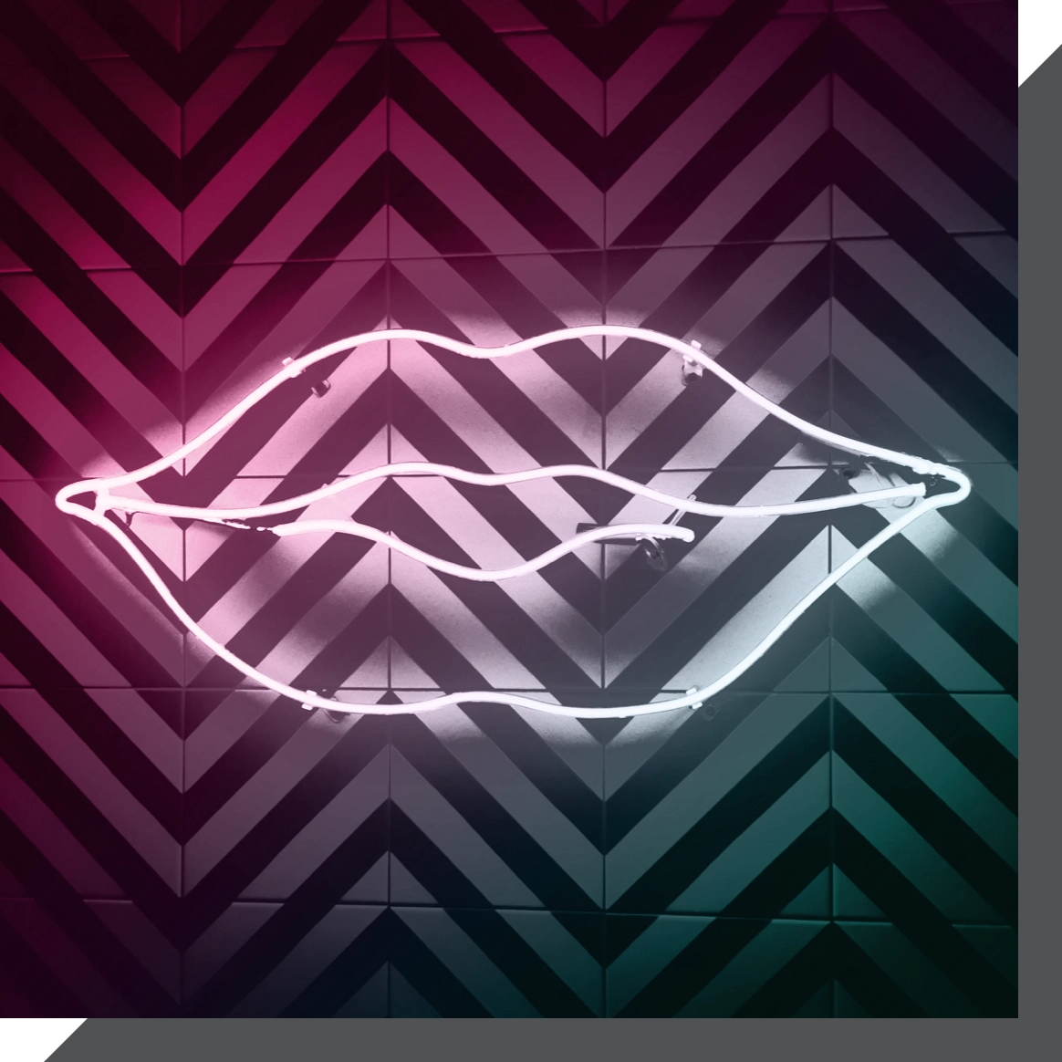 A neon light sculpture in the shape of lips is mounted on a chevron-patterned wall. The wall features a gradient of dark pink and teal colors. The neon light is white and is the focal point of the image.