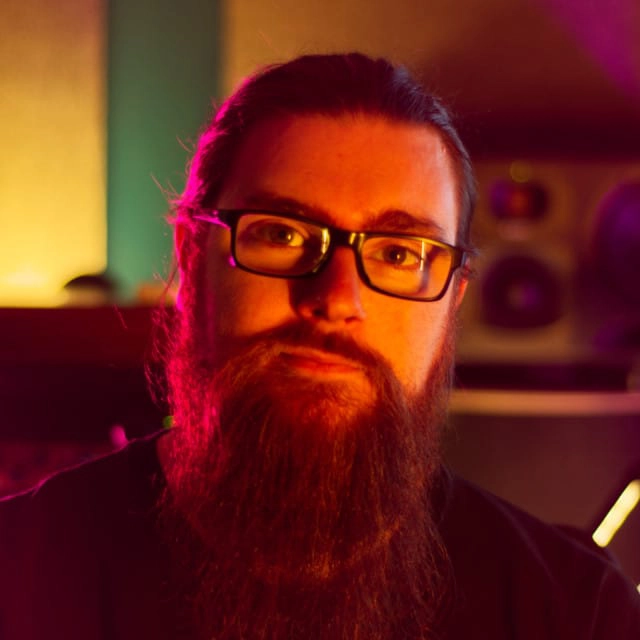 A person with glasses and a long beard is looking at the camera. The background features various colored lighting, giving the image an atmospheric feel. There are speakers and what appears to be musical equipment visible behind the person.