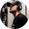 A man wearing a black cap and headphones is singing into a microphone in what appears to be a recording studio in London. He is dressed in a black shirt and looking to the side, concentrating on his performance.