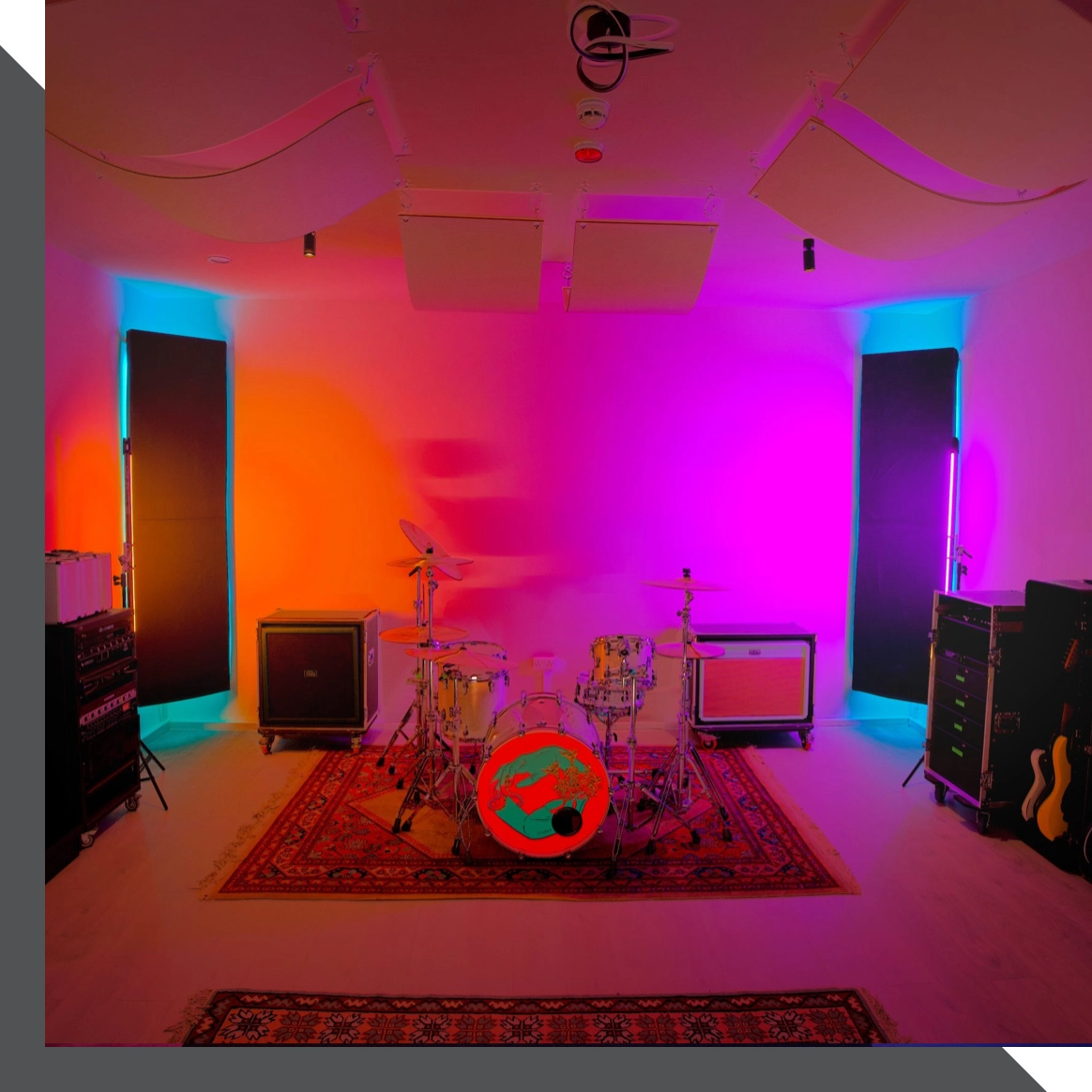 A recording studio in London featuring a drum set with a painted bass drum head at the center, surrounded by amplifiers and sound equipment. The room is illuminated by vibrant colored lights, creating a gradient effect on the walls. Guitars are positioned to the right.