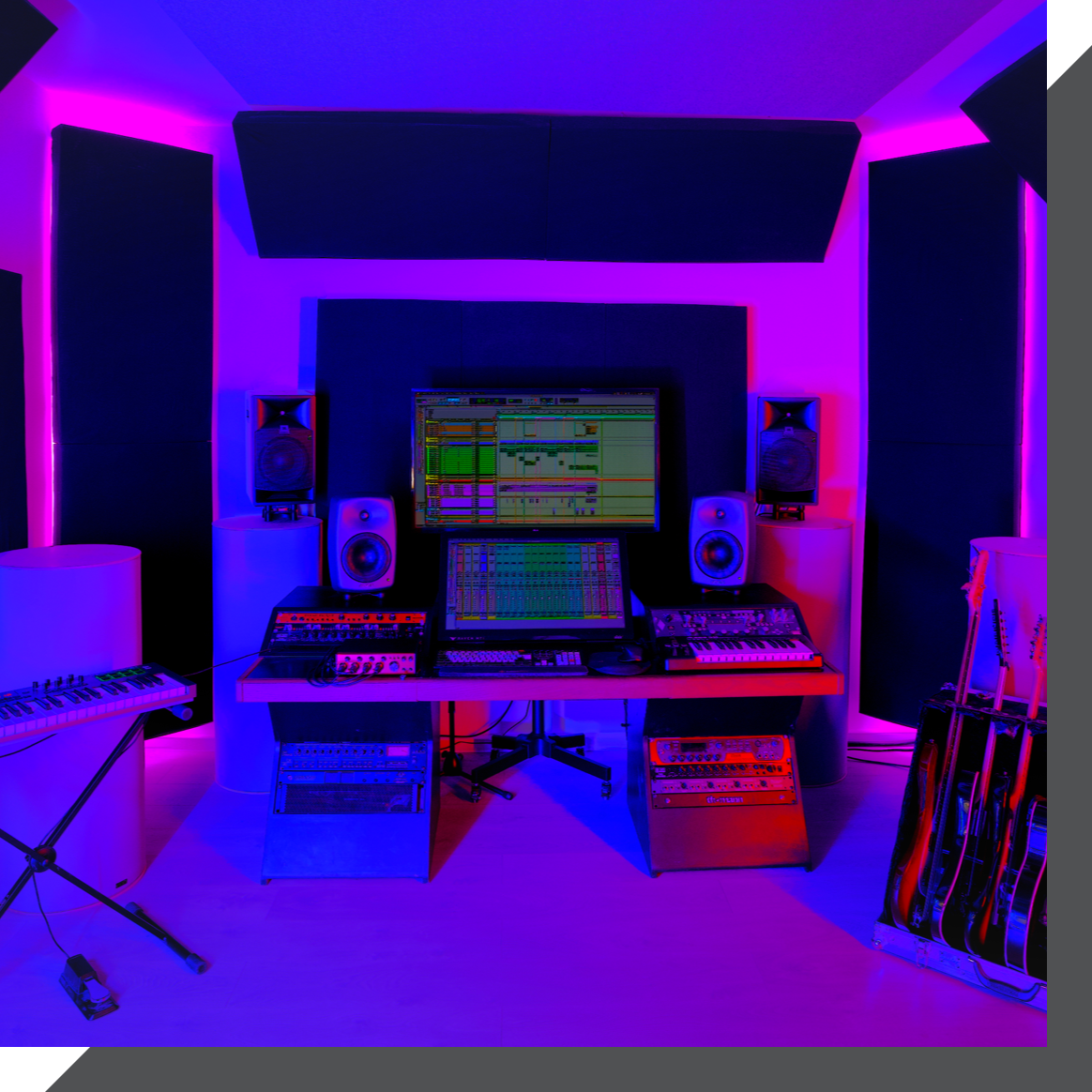 A recording studio in London with a central computer screen displaying audio software, surrounded by speakers, various keyboards, mixers, and six guitars standing in a rack. The room is illuminated with purple and blue lighting, creating a vibrant atmosphere.