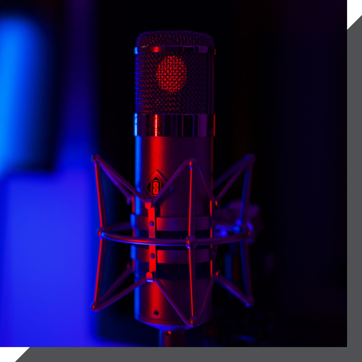 A close-up shot of a studio microphone illuminated by red and blue lights in a recording studio in London. The microphone is mounted on a shock mount, and the background is out of focus with a mix of blue and purple hues.