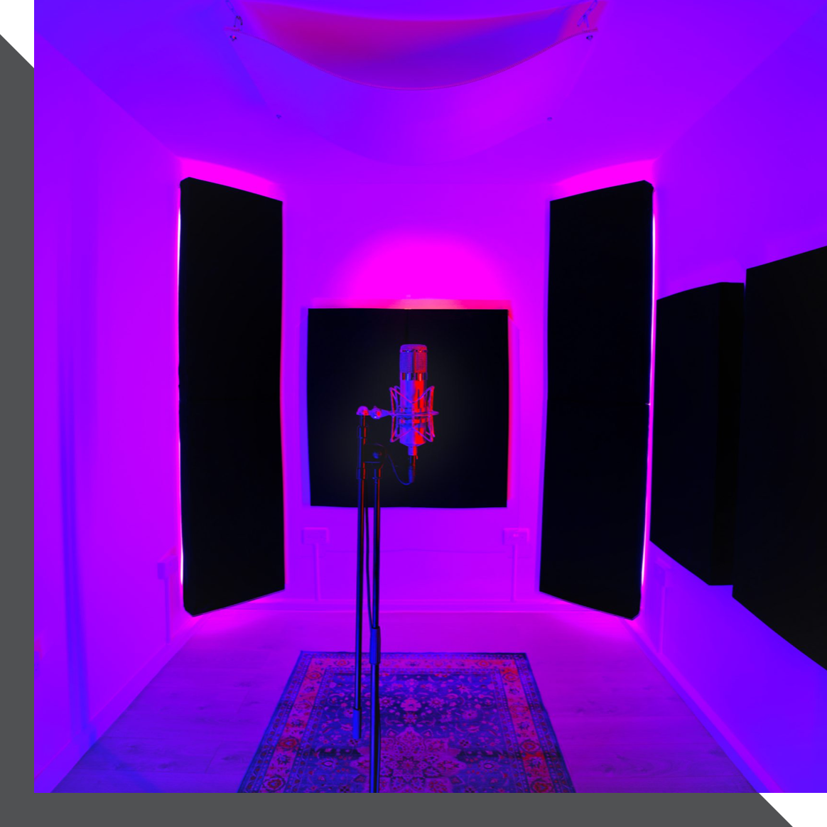 A recording studio in London is illuminated by purple and pink lights. A microphone is set up on a stand in the center of the room, which has white walls and acoustic panels. A patterned rug lies on the floor beneath the microphone.