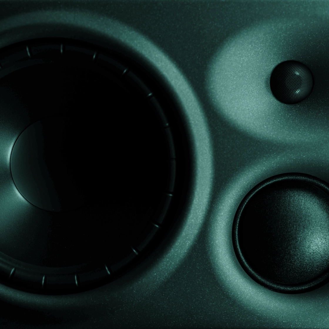 Close-up image of a speaker system featuring a large central woofer, a smaller subwoofer to the bottom right, and a small tweeter to the top right, all housed in a dark, textured casing. The lighting creates subtle reflections and shadows on the surface.