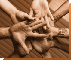 Six hands, all with palms facing upwards, are overlapping each other in a circular formation. The image is taken in a sepia tone. One of the hands has a tattoo on the forearm with visible text. The background, reminiscent of a recording studio in London, consists of wooden planks.