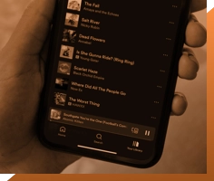 A hand holding a smartphone displays a music playlist. The screen shows a list of songs, with the currently playing track being "Sunflower You’re the One (Product’s Comet Remix)" by Product. The background, suggestive of a recording studio in London, is out of focus.