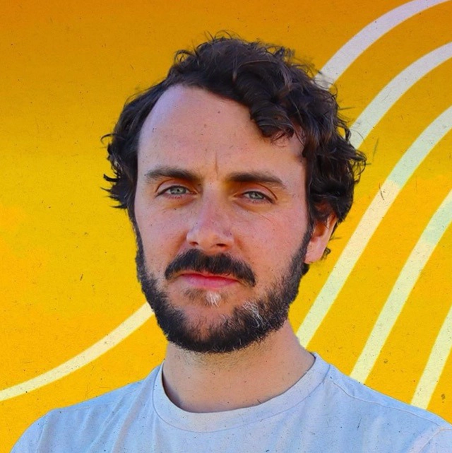 A man with curly dark hair and a beard stands in front of a vibrant yellow background with white curved lines, evoking the creative vibe of a recording studio in London. He is wearing a light-colored shirt and looking directly at the camera.